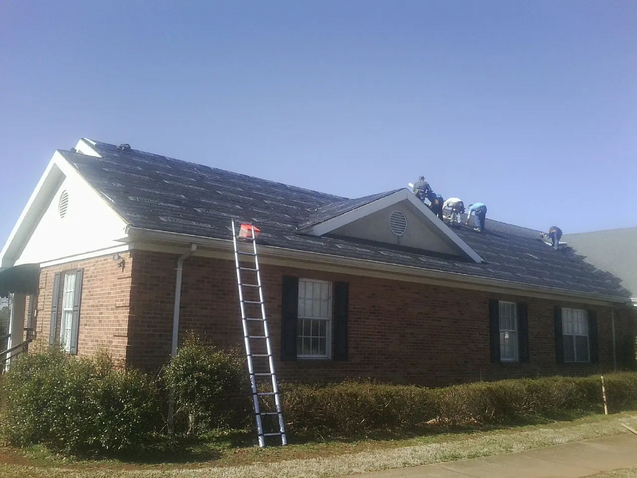 Crew tearing shingles off a brick ranch roof with a ladder set — Metal Roof Repair work by our crew in Herman